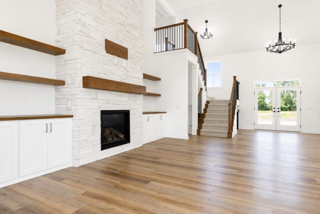 Spacious great room with a floor-to-ceiling stone fireplace, floating wood shelves, custom built-in cabinetry, open staircase, luxury vinyl plank flooring, and abundant natural light in a Columbia City, Indiana custom home by Wannemacher Design Build.