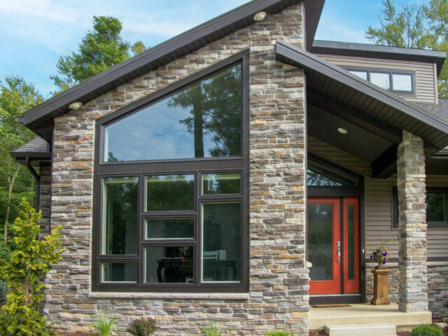 Custom home exterior featuring stone façade, expansive geometric window, and covered entry with modern red door.