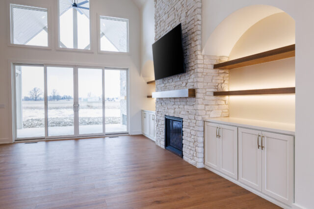 Custom home great room in Fort Wayne with stone fireplace, built-in shelving, and large sliding glass doors overlooking the backyard.