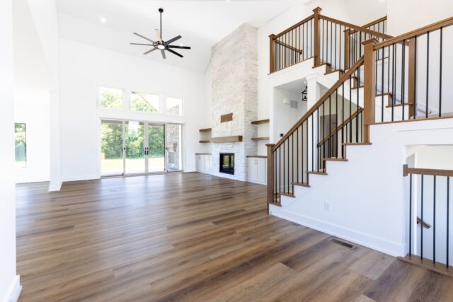 Open-concept great room featuring a floor-to-ceiling stone fireplace, custom staircase with wood and iron railing, expansive windows, luxury vinyl plank flooring, and covered patio access in a Columbia City, Indiana custom home by Wannemacher Design Build.